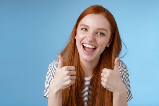 Enthusiastic amused outgoing redhead beautiful girl say yeah supportive like awesome idea show thumbs-up approval recommend gesture agree good choice, standing blue background. Copy space