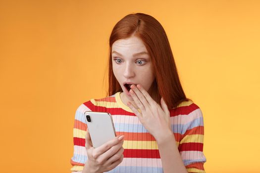 Lifestyle. Amazed speechless young teenage redhead girl student gasping drop jaw say omg wow cover opened mouth palm look shocked surprised smartphone display reading fresh gossips orange background.