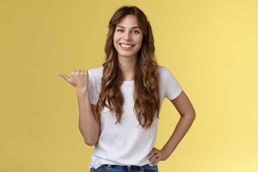 Friendly outgoing cheerful european cute female giving tips showing direction lively smiling toothy happily have nice pleasant conversation pointing left thumb introduce promo yellow background.