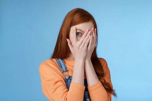 Not peeking promise. Charming intrigued cute redhead teenage girl hiding face cover eyes palms look through fingers check out gift anticipating something interesting standing curious blue background.