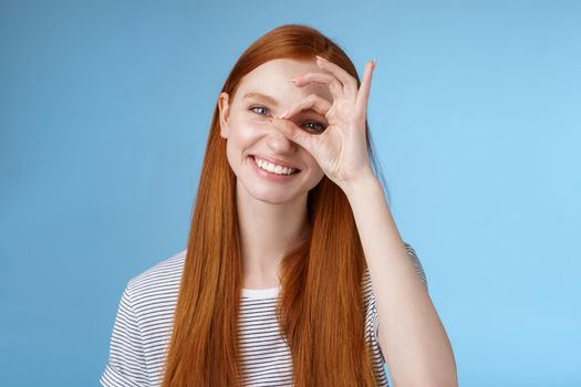 Charismatic happy adorable redhead teenage girl sincere eyes making circle eye show okay ok sign delighted like approve cool idea smiling satisfied achieve perfect score, standing blue background.