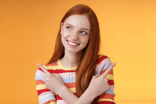 Dreamy cute redhead girl planning were go summer holidays have different choices picking variant look intrigued smiling pleased pointing sideways left right taking decision, orange background.