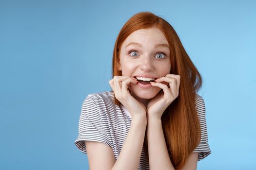 Excited amazed attractive redhead girl blue eyes look fascinated affection smiling desire bite fingernails eager feel astonished cannot wait bite tasty food, standing blue background thrilled.