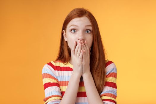 Shocked speechless impressed sensitive redhead european girl reacting stunning rumor gossiping find out secret gasping cover mouth palm stare camera astonished surprised, orange background.