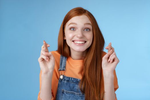 Excited emotional happy cheerful redhead girl smiling optimistic stare surprised thrilled cross fingers good luck believe dream come true make wish anticipating only good news, blue background.