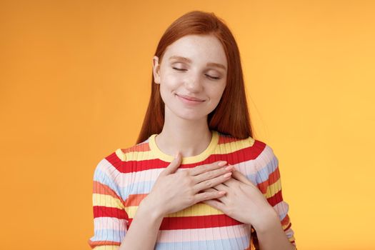 Heart keeps warm save dearest memories inside. Smiling tender heartwarming redhead lovely girl touch chest grinning closed eyes recall lovely nice moment standing romantic orange background.