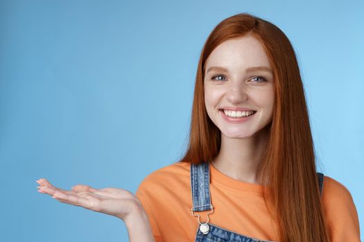 Proud good-looking confident redhead girl present awesome product hold object palm raise hand blank blue copy space smiling delighted recommend cool link, standing studio background helpful.