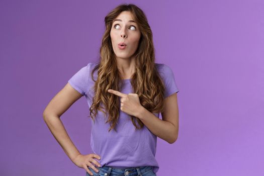 Intrigued lively attractive feminine curly-haired female pointing looking left impressed folding lips say wow astonished express admiration enthusiastic check out awesome promo purple background.