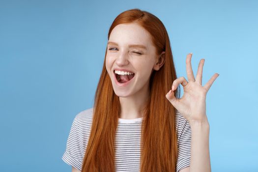 Sassy attractive redhead girl winking mysteriously smiling broadly give approval sign show okay ok excellent gesture satisfied good choice agree great decision, standing delighted blue background.