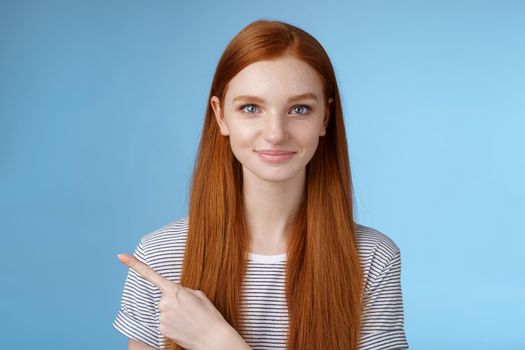 Attractive modest good-looking redhead girlfriend acting mature friendly smiling pointing left index finger giving direction show way bathroom standing blue background joyful kind grin.