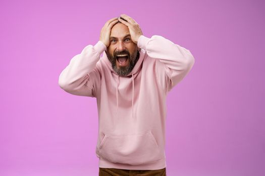 Outraged furious mad adult man bearded yelling shocked despair hatefully looking camera insane holding hands head frowning shouting losing bet disappointed very upset standing purple background. Lifestyle.