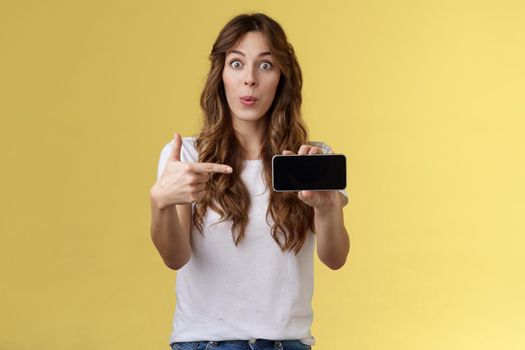 Excited surprised pleased girl curly long hairstyle folding lips whistling amused stare camera impressed showing smartphone pointing index finger mobile cellphone screen stand yellow background.