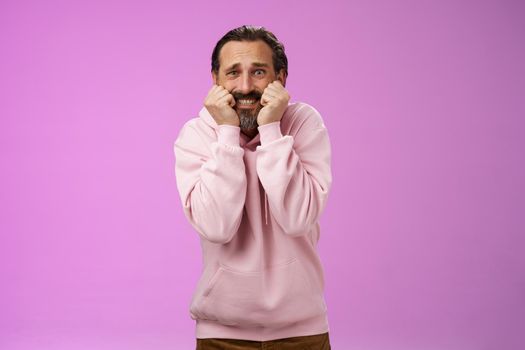 Scared insecure silly adult bearded man grey hair in pink hoodie press palms mouth bite fingers clench teeth shocked frightened widen eyes terrified standing stupor horrified, purple background.