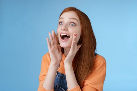 Attractive silly european redhead young girl 20s calling friend searching someone crowd look relaxed joyfully yelling hold hands opened mouth shouting name louder look left, blue background.