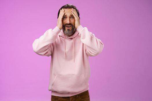 Adult upset bearded man despair standing troubled concerned touching face frowning grimacing sorrow unhappiness, losing bet, was robbed, standing shocked devastated purple background, panic.
