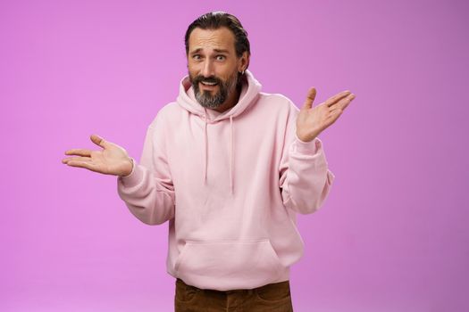So what bite me. Portrait ignorant careless cool stylish mature bearded man earring pink hoodie shrugging hands sideways mocking being rude standing pissed unwilling help standing purple background.