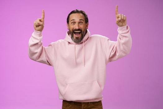 Energized happy good-looking trendy mature dad beard grey hair in pink stylish hoodie wearing son outfit pointing up index fingers thrilled favorite music group in town, standing purple background.