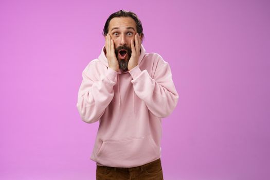 Amused very happy adult bearded dad receive incredible impressive bunch money screaming joyfully touch cheeks widen eyes astonished cannot believe how lucky, standing purple background thrilled.