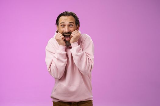 Amused thrilled gasping handsome happy bearded adult man in pink hoodie stooping screaming joyfully receive awesome incredible present standing speechless excited purple background. Copy space