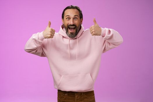 Excited energized supportive charismatic european mature bearded man in pink hoodie show thumbs up gesture smiling agree approving recommending awesome product, standing purple background satisfied.