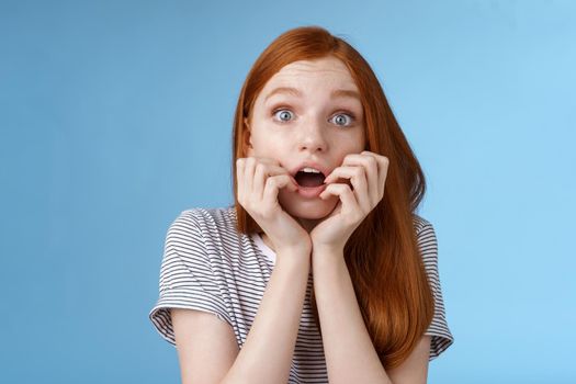 Shocked speechless gasping young redhead girl staring impressed stunned watching important moment tv series biting fingers open mouth shook standing excited blue background anticipating.