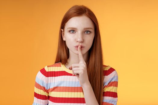 Girl hides lots secrets. Serious-looking redhead girlfriend blue eyes demand keep silent voice down show shhh shush gesture index finger pressed lips, prepare surprise, orange background.