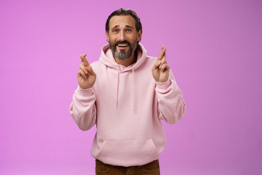 Hopeful lucky optimistic worried mature stylish bearded man grey hair in trendy hoodie cross fingers wish anticipating important results nervously standing purple background frowning anxious.