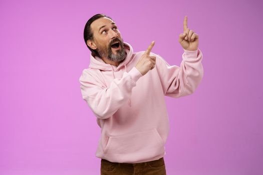 Enthusiastic energized charismatic grandfather bearded grey hair in hipster modern hoodie pointing looking impressed fascinated upper right corner discussing interesting fashion trends.