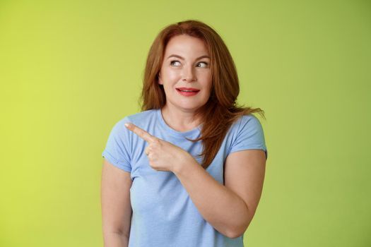 Excited intrigued middle-aged mature redhead woman. pointing gazing left side copy space curiously smiling thrilled like interesting promo willing check-out great advertisement green background.