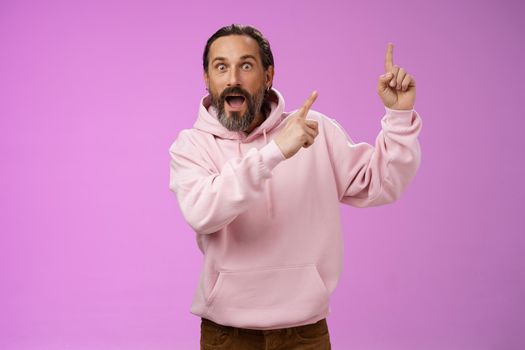 Impressed shocked exctied cool hipster mature 50s man bearded grey hair in pink hoodie retelling incredible story pointing up index fingers widen eyes surprised thrilled awesome advertisement. Lifestyle.