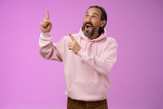 Fashionable good-looking fascinated mature bearded man grey hair look astonished pointing gazing upper left corner see miracle incredible breathtaking magnificanent view, standing purple background.