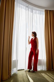 Elegant young curly woman standing next to the curtains on big panoramic window