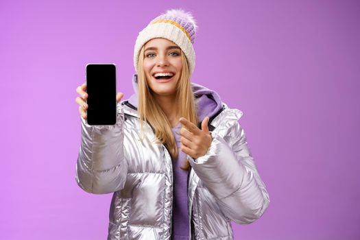 Satisfied amused good-looking blond girl suggest take look smartphone display smiling happily pointing mobile phone delighted talking about awesome new app features, standing purple background.