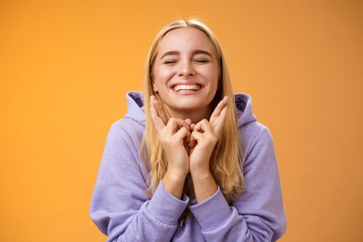 Charming lucky cute young happy hopeful blond girl close eyes smiling positive cross fingers good luck wanna win faithfully praying make wish desire dream come true, standing orange background.