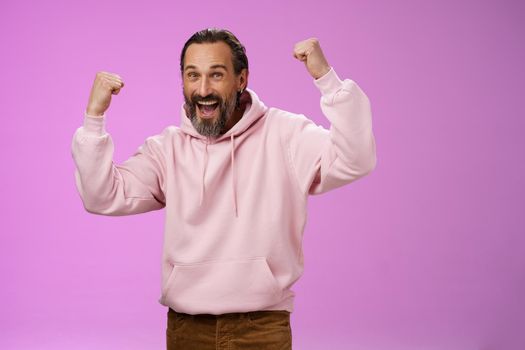 Yeah who cares age. Carefree delighted happy old man bearded in trendy pink hoodie raise fists joyfully triumphing having fun glad win celebrating success accomplish goal, posing purple background.