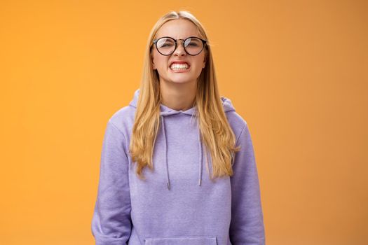 Blond girl show teeth warn can stand herself bite, fool around grimacing funny wearing purple hoodie glasses standing orange background better not mess with me, pranking friend have fun.