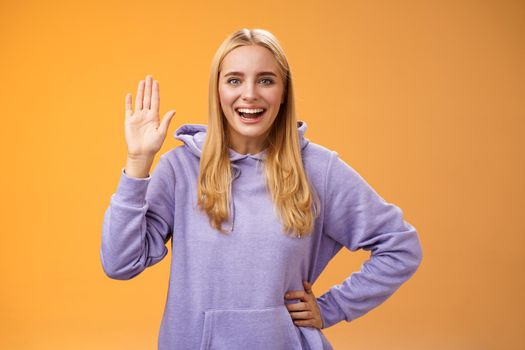 Friendly energized helpful charming female student say hello newcomers waving raised palm hey hi gesture greeting welcoming happily, standing orange background smiling broadly. Copy space