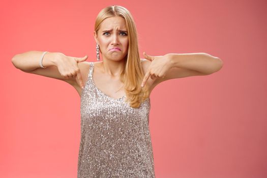 Whining complaining upset depressed blond woman in silver dress crying boyfriend broke-up prom night standing unhappy expressing sorrow regret disappointment pointing down red background.