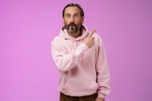 Amused handsome hipster mature 40s bearded caucasian man grey hair wearing trendy pink hoodie fold lips amazed wow pointing upper right corner widen eyes surprised check out awesome product. Lifestyle.