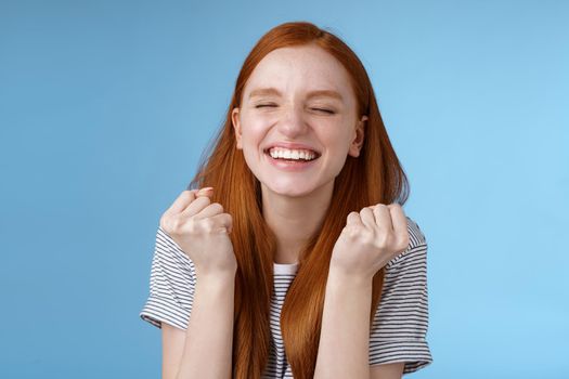 Lifestyle. Happy accomplished good-looking ginger girl receive positive results test clench fists rejoicing yelling yes celebrating do winner dance close eyes relieved standing blue background triumphing.