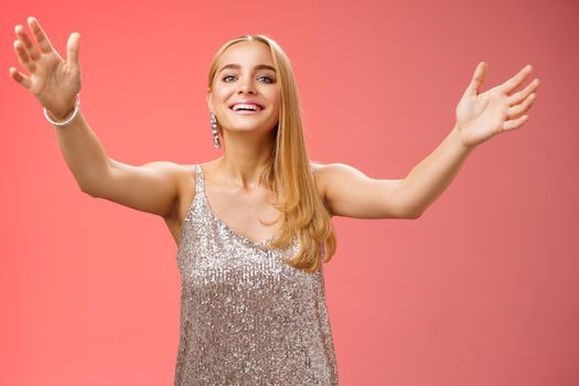 Friendly charming tender hospitable attractive birthday girl in luxury silver dress welcoming guests throw party extend arms cuddle greeting people hug give cuddle smiling broadly, red background.