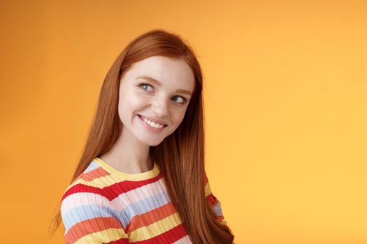 Silly flirty good-looking redhead girl making first move checking out guy standing profile turning left smiling intrigued glancing aside coquettish flirting seducing sassy, orange background.