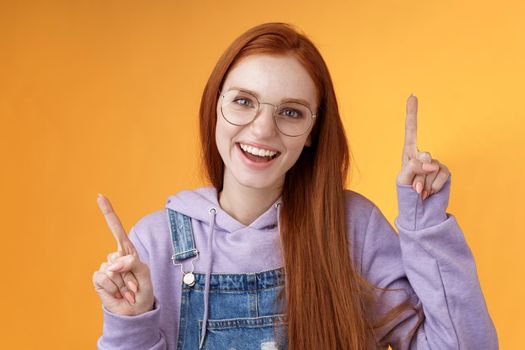 Lively enthusiastic millennial redhead girl coworker having fun celebrating small break joyfully dancing pointing up index fingers singing smiling white teeth promoting product, orange background.