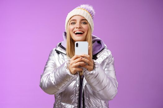 Amused excited attractive blond girlfriend holding smartphone up recording video boyfriend step snowboard first time capturing memories mobile camera standing happily purple background.