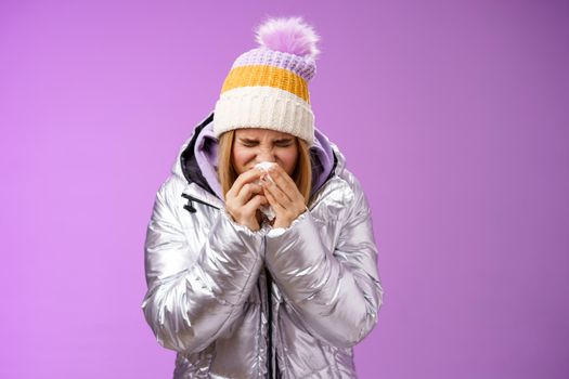 Girl runny nose sneeze tissue press napkin face got ill feeling unwell sick heading hospital standing purple background bending wearing silver stylish jacket winter hat, crying sobbing.