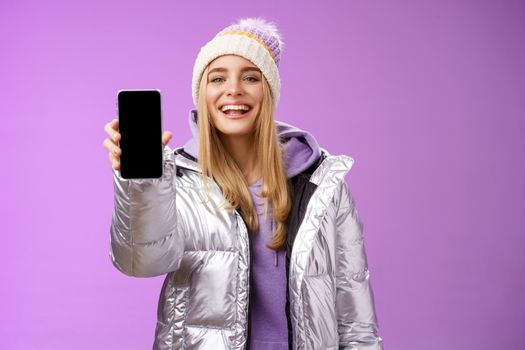 Sassy delighted cheerful blond woman recommend use app edit perfect pictures holding smartphone showing mobile phone display proudly satisfactory smile camera, standing purple background.