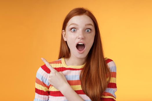 Impressed speechless young redhead 20s girl drop jaw gasping amused wide eyes staring camera surprised see awesome stunning new product pointing left index finger questioned, orange background.