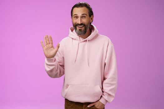 Cheeky charismatic funny happy smiling mature man bearded grey hair in pink hoodie waving palm hello nice meet you greetng gesture welcoming guests say hi standing purple background friendly relaxed. Lifestyle.