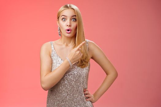 Wondered amused attractive blond woman talking party widen eyes say wow pointing questioned right direction see famous person standing surprised impressed gaze camera, red background.