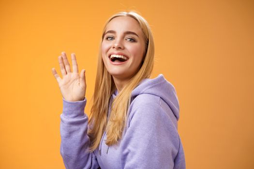 Waist-up friendly lucky cute blond girl waving palm happily smiling turning camera greet friend say hello hi see familiar person welcoming newcomers joyfully, standing orange background.